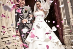 mariée le bras en l'air tenant son bouquet au bras de son mari à la sortie de l'église sous une pluie de pétales blancs et roses