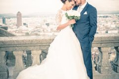 jeune mariée à la longue robe et au bouquet de roses blanches enlacée dans les bras de son mari sur le bord de la terrasse de la basilique de Fourvière surplombant Lyon