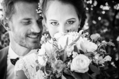 jeune mariée au regard pénétrant sourie cachée derrière son bouquet avec son mari qui lui sourit