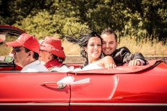 couple de jeunes mariés sourit les cheveux au vent à l'arrière d'un cabriolet rouge