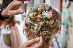 mains d'une coiffeuse finalisant le chignon d'une future mariée avec des roses dans ses cheveux blonds