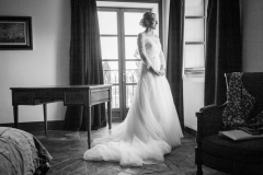 future mariée blonde en robe blanche à longue traine regardant par la fenêtre de la chambre entourée de vieux meubles en bois