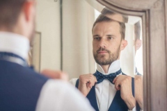 reflet du futur marié blond et barbu ajustant son noeud papillon assorti à son costume trois pièces bleu pendant ses préparatifs