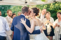 couple de mariés s'embrasse sous une tonnelle pendant le cocktail de leur mariage entouré de leurs invités